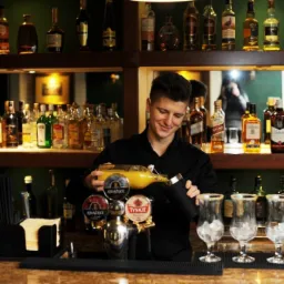 Barman w czarnej koszuli przygotowuje drinka, nalewając żółty płyn z butelki do shakera. W tle półki z alkoholem, firmowe nalewaki i szklanki z lodem.