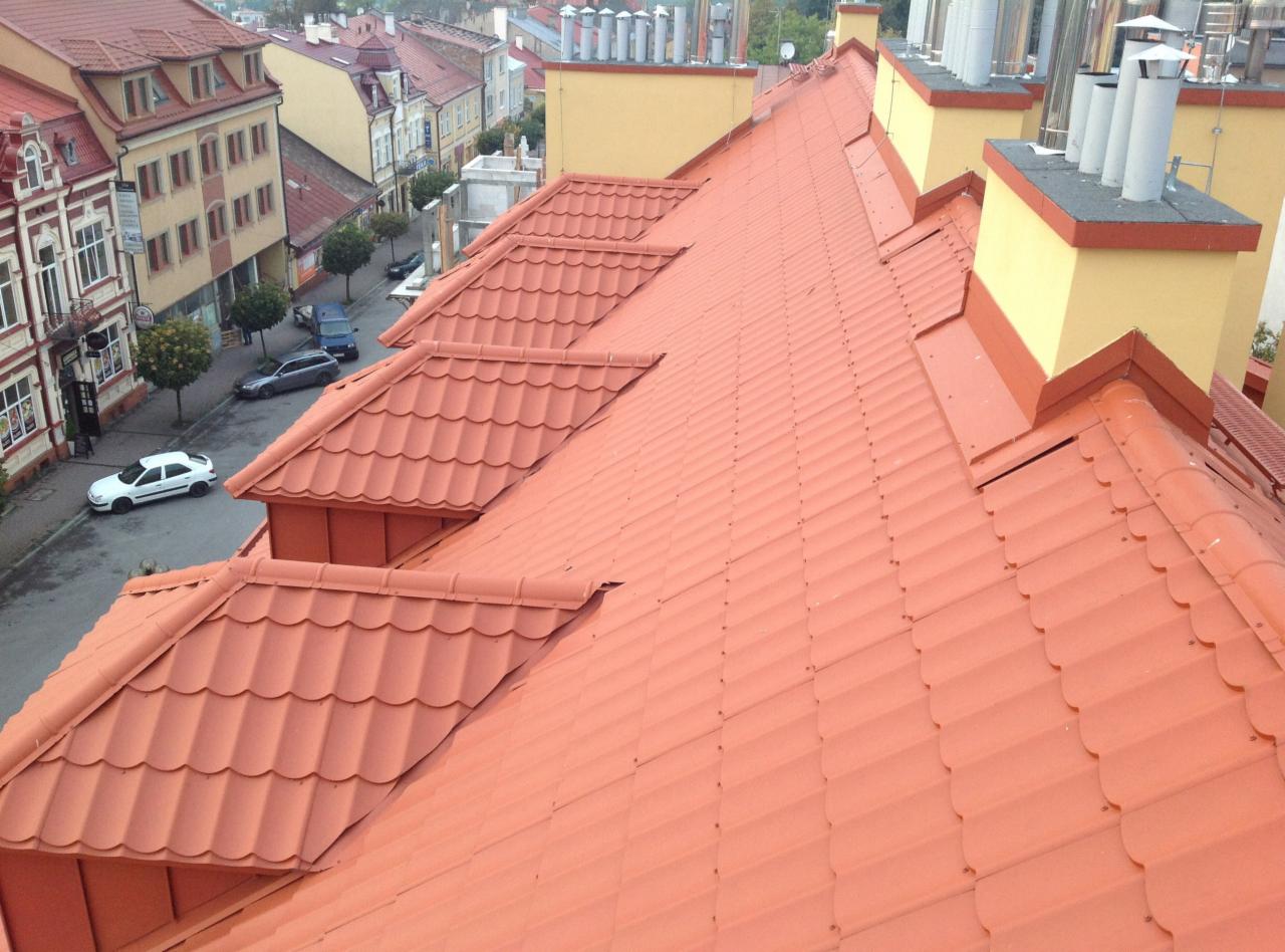 Czerwony dach z blachodachówki z widokiem na miasto, kominy w tle. Układ dachówek w regularnych rzędach. Perspektywa z góry, ukazująca detale pokrycia.