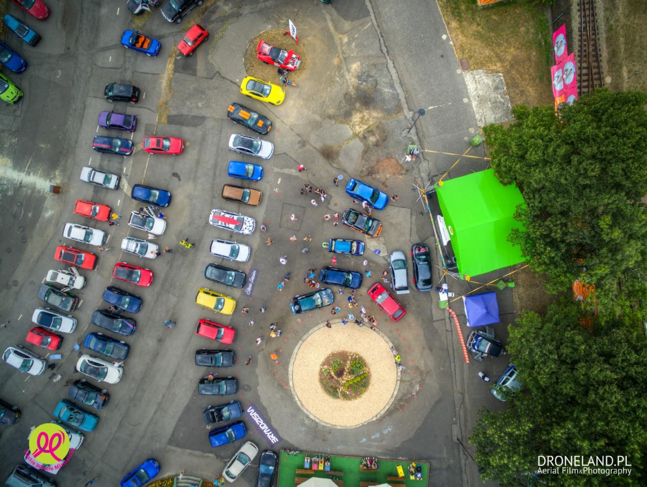 Widok z drona na parking pełen kolorowych samochodów podczas zlotu. Uczestnicy spacerują, a w tle widoczny zielony namiot i drzewa. Zdjęcie z lotu ptaka.