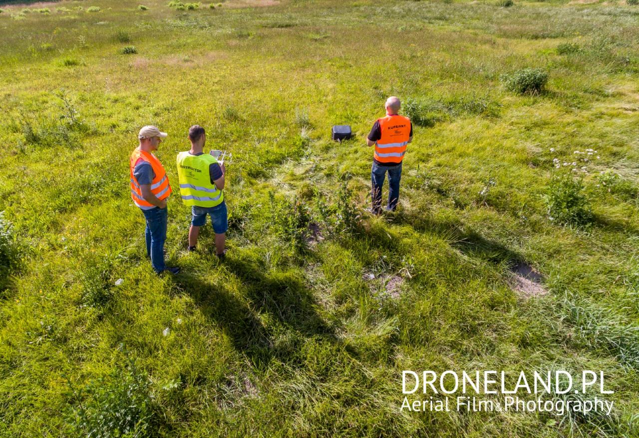 Z lotu ptaka: Trzech mężczyzn w kamizelkach odblaskowych (dwóch z napisem 'Instruktor', jeden 'Kursant') obsługuje drona na łące.