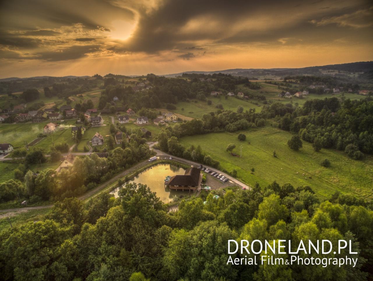 Panoramiczny widok z drona na karczmę nad wodą o zachodzie słońca, otoczoną zielonymi wzgórzami i polami, z widocznymi domami w oddali.