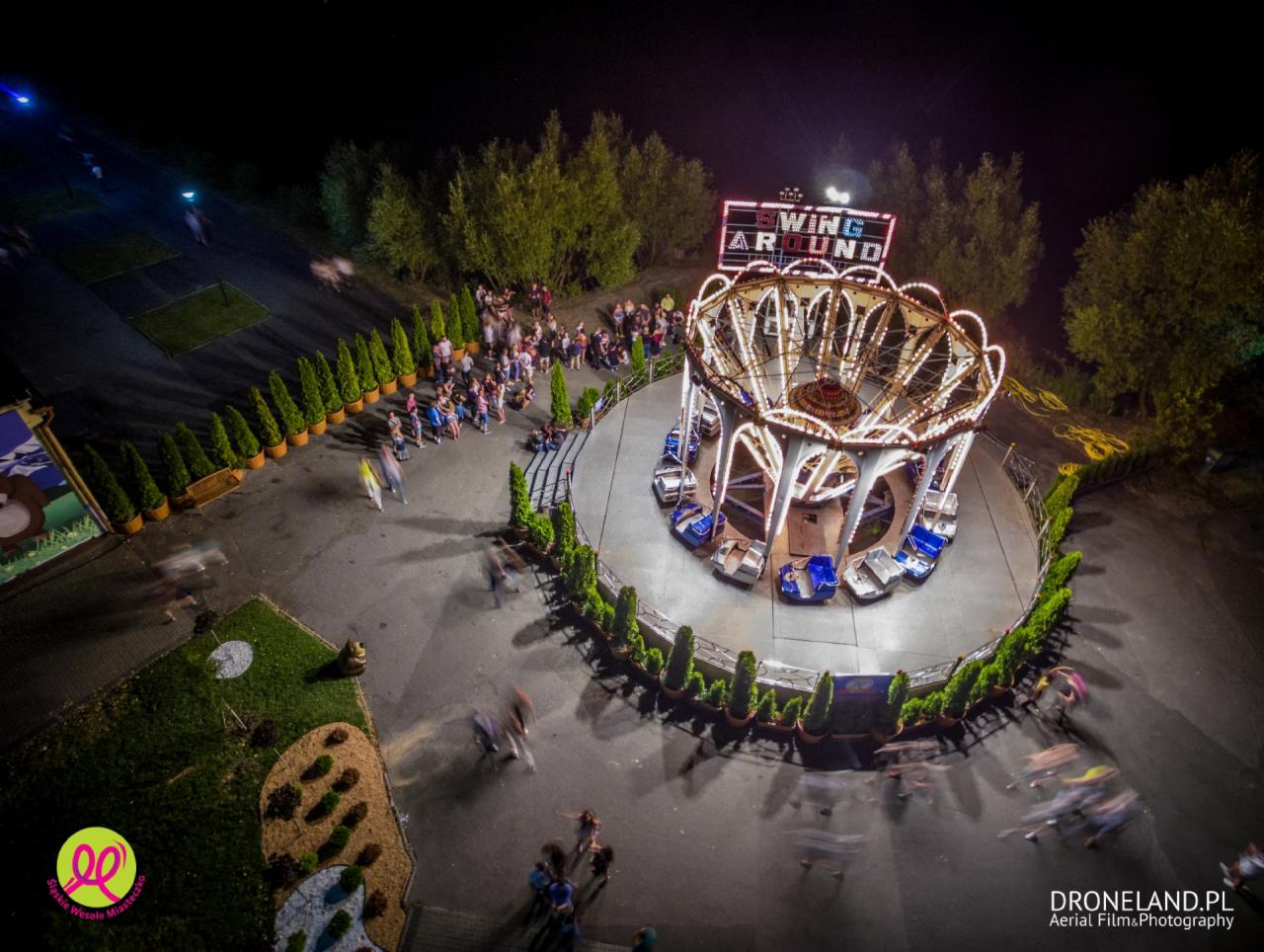Nocne ujęcie z drona na karuzelę łańcuchową 'Wing Around' w Wesołym Miasteczku w Chorzowie, otoczoną tłumem ludzi i zielenią, z widocznym oświetleniem i rozmytymi postaciami w ruchu.