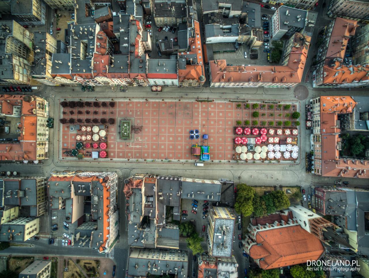 Widok z lotu ptaka na Rynek w Bytomiu, prezentujący geometryczny układ placu z czerwonej kostki, otoczonego budynkami z czerwonymi dachami. W centralnej części placu widoczna fontanna i rzędy drzew...