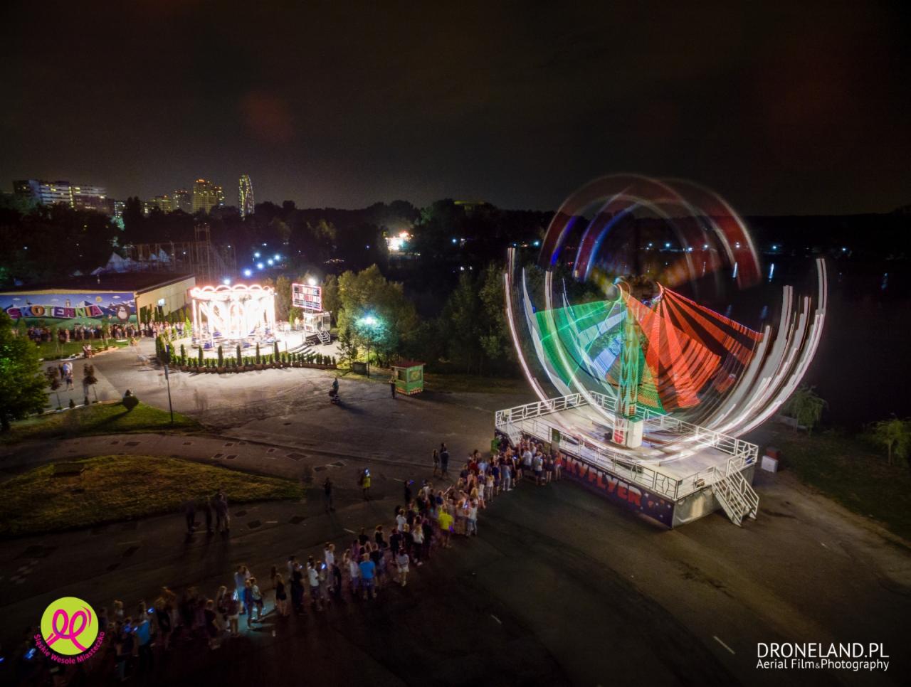 Nocne ujęcie z lotu ptaka na wesołe miasteczko z karuzelą i kolejką ludzi, w tle budynki miejskie.