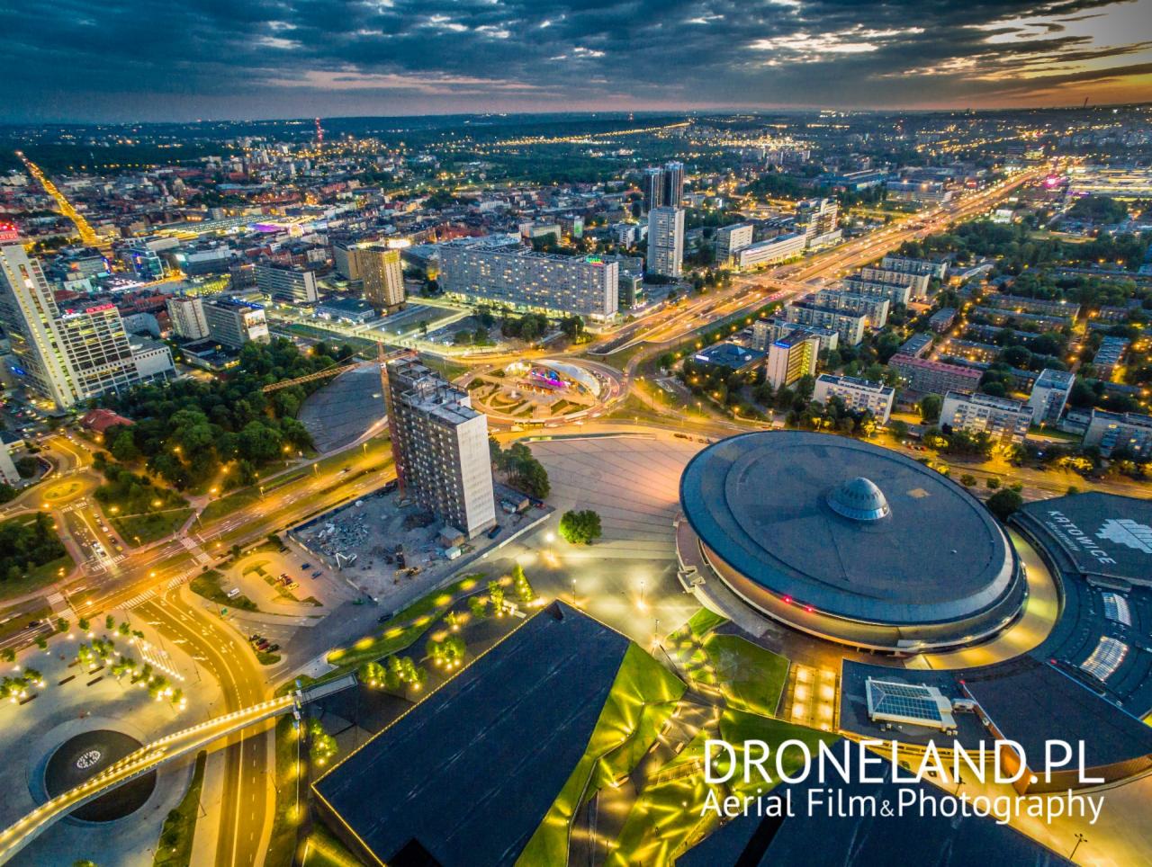 Nocna panorama Katowic z lotu ptaka, widoczne Spodek, budynki, ulice oświetlone światłami, wieczorne niebo z chmurami, logo Droneland.pl w prawym dolnym rogu.
