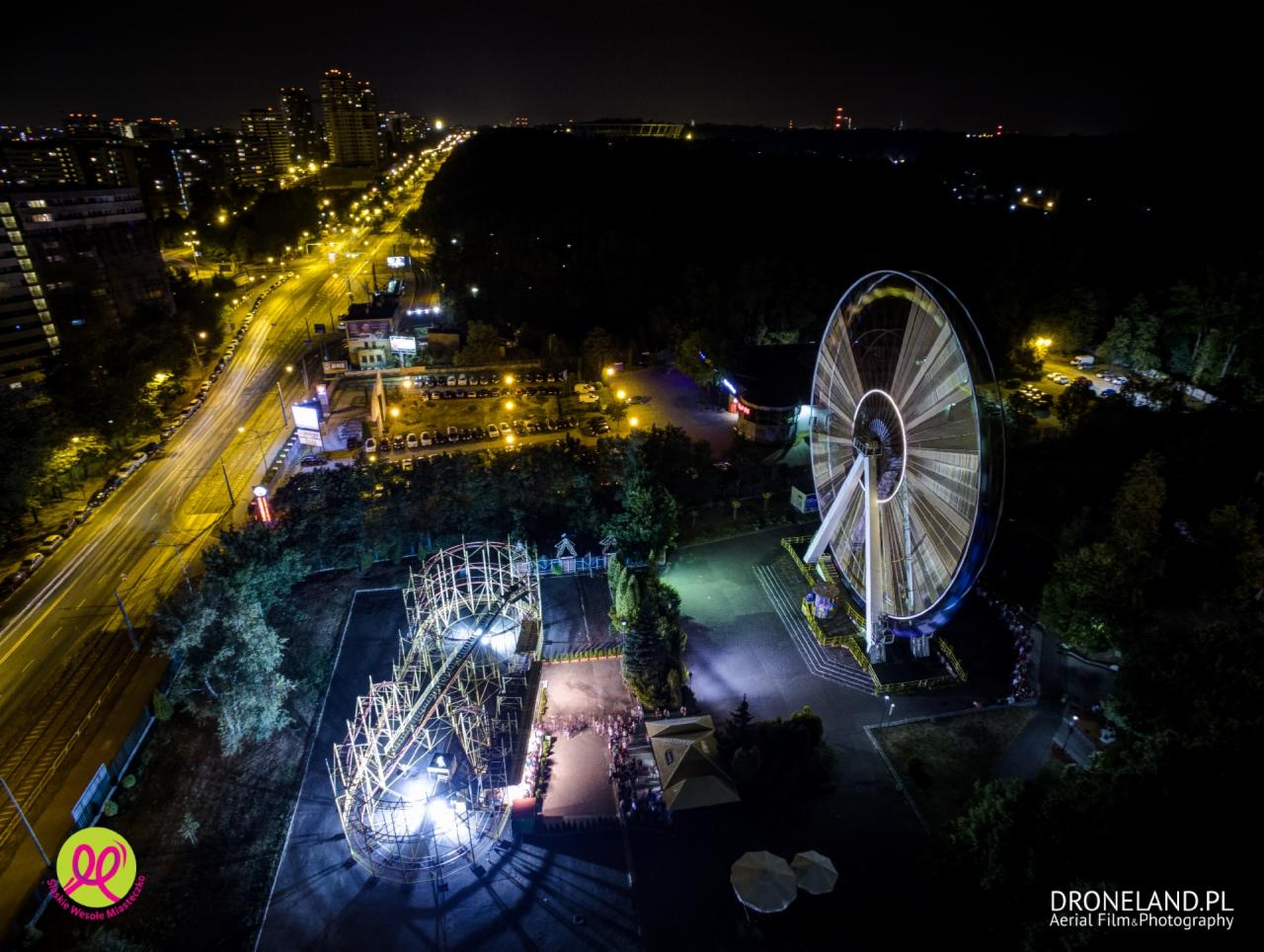 Nocny widok z lotu ptaka na oświetlone diabelskie koło i kolejkę górską w Wesołym Miasteczku Magicland, z widoczną panoramą miasta w tle.