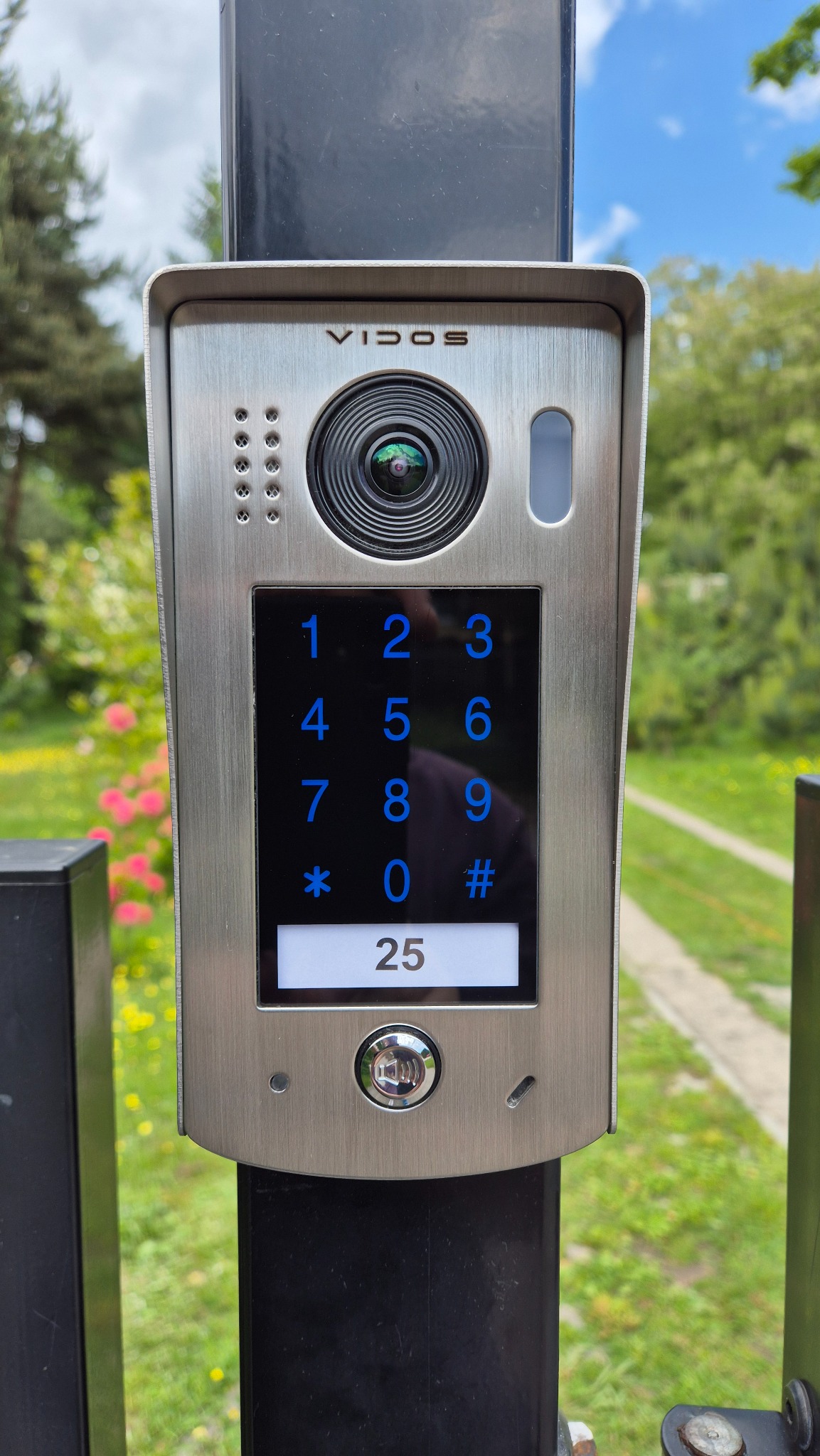 Nowoczesny wideodomofon Vidos z podświetlaną klawiaturą numeryczną i kamerą, zamontowany na czarnym słupku na tle zieleni.