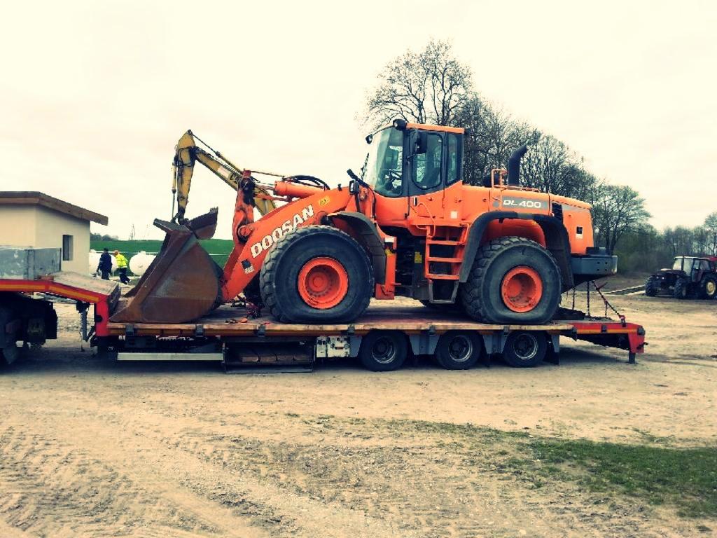 Pomarańczowa ładowarka Doosan DL400 na niskopodwoziowej lawecie, zabezpieczona łańcuchami, w tle budynek i drzewa. Transport maszyny budowlanej.