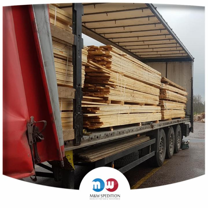 Ciężarówka z czerwoną plandeką załadowana drewnianymi deskami na paletach, widok z boku, logo firmy transportowej w rogu zdjęcia.