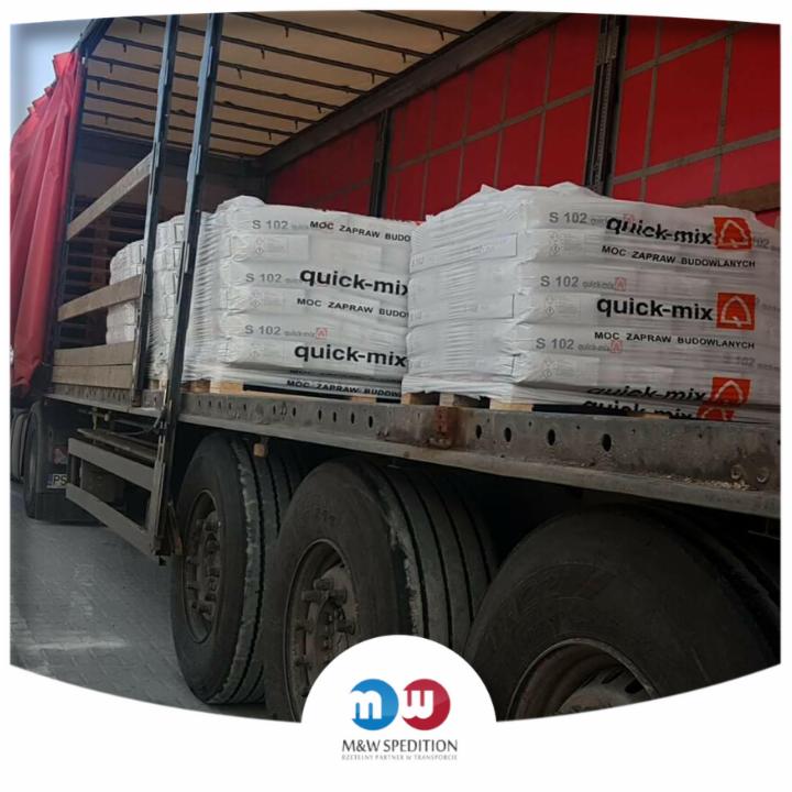 Ciężarówka z czerwoną plandeką przewożąca palety z workami zaprawy budowlanej Quick-Mix S102, widok z boku na koła i część ładunku.