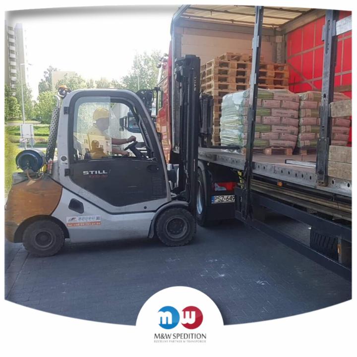 Wózek widłowy STILL ładuje palety i worki z materiałami budowlanymi do naczepy ciężarówki, widoczny operator w kasku, tablica rejestracyjna pojazdu PSZ 649, w tle budynek mieszkalny i zieleń.