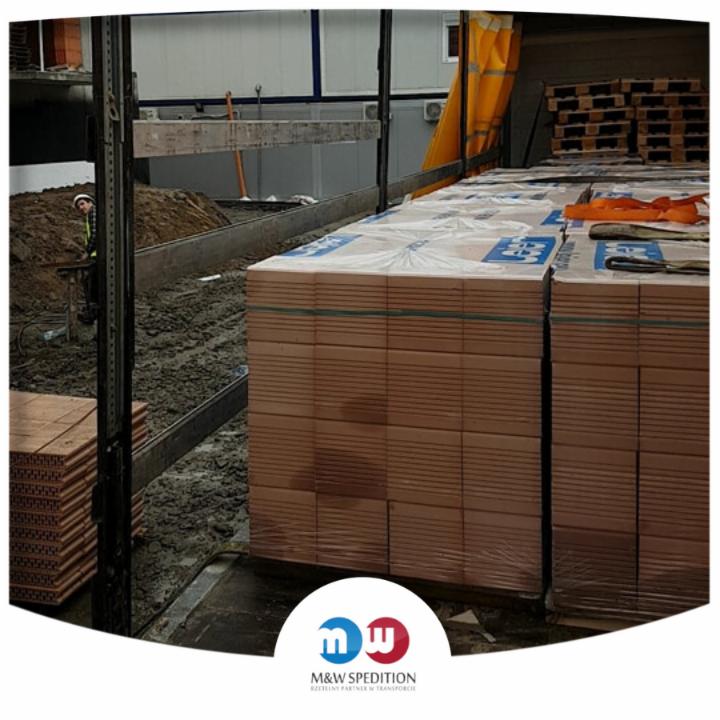 Cegły ceramiczne zapakowane w folię, zabezpieczone pasami, załadowane na naczepie ciężarówki, w tle widoczny pracownik w kasku.