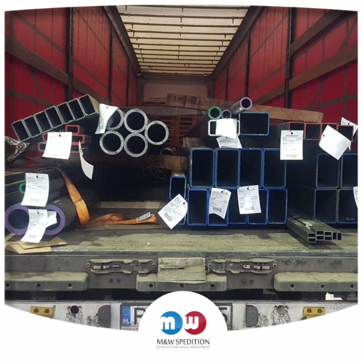 Wnętrze naczepy wypełnione różnymi profilami stalowymi, rurami okrągłymi i kwadratowymi, zabezpieczonymi pasami transportowymi, z widocznymi etykietami adresowymi.