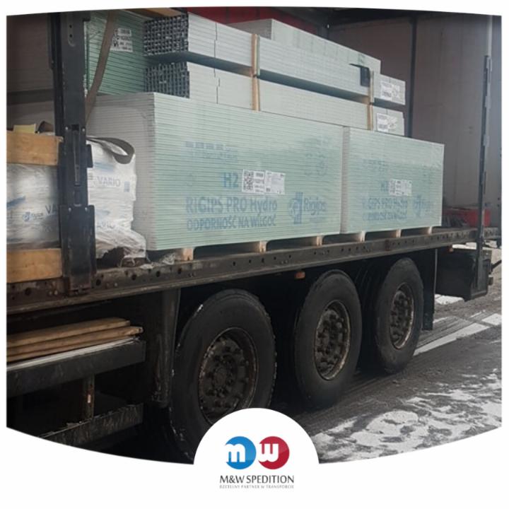 Ciężarówka z naczepą przewozi palety z płytami gipsowo-kartonowymi Rigips Pro Hydro odpornymi na wilgoć, oraz profile metalowe, widoczne opony i części podwozia.