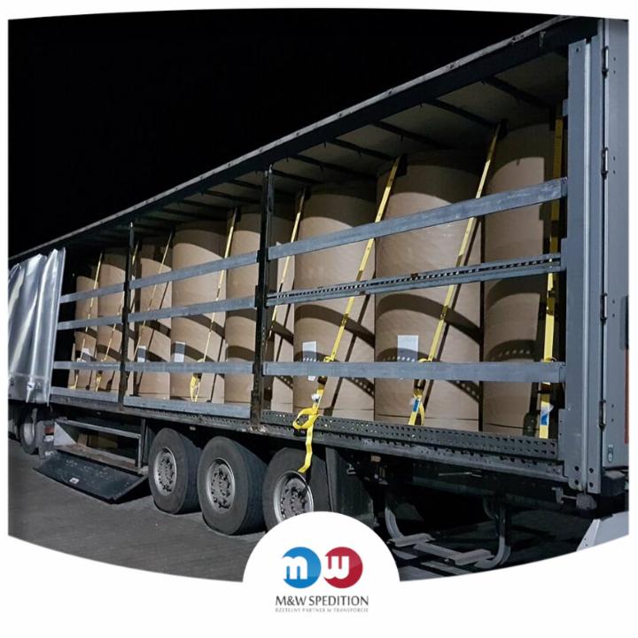 Ciężarówka z otwartą naczepą, załadowana rolami papieru zabezpieczonymi żółtymi pasami, widoczna w nocy.