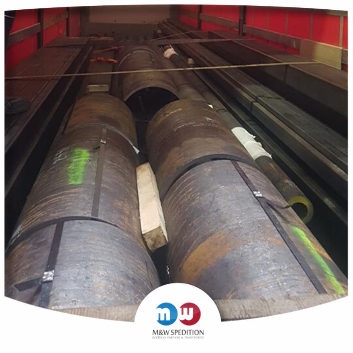 Cylindryczne metalowe ładunki zabezpieczone pasami transportowymi wewnątrz naczepy, widoczne również profile stalowe i fragment czerwonej plandeki.