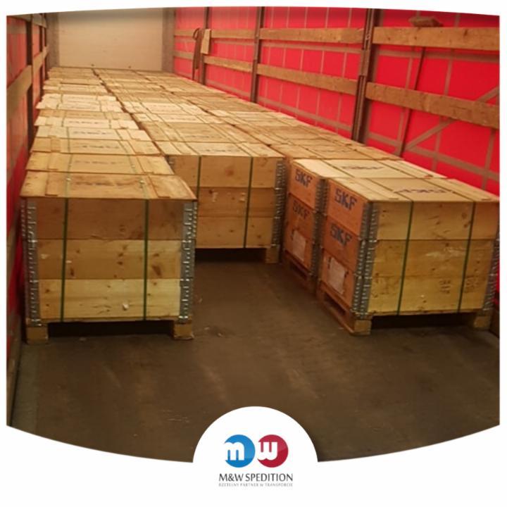 Wnętrze przestrzeni ładunkowej wypełnione drewnianymi skrzyniami SKF na paletach, zabezpieczonymi zielonymi taśmami, widoczne czerwone ściany pojazdu.