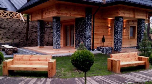 Dwie drewniane ławki ogrodowe przed domem z bali z kolumnami obłożonymi kamieniem naturalnym.