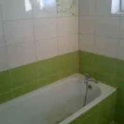 Biało-zielona łazienka po remoncie, widoczna wanna obłożona zielonymi płytkami, ściany wyłożone białymi płytkami z delikatnym wzorem. Ujęcie z rogu łazienki.