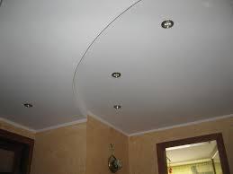 Biały, podwieszany sufit z oświetleniem punktowym w narożnym wnętrzu. Listwa dekoracyjna przy ścianie. Ściany w odcieniach beżu i brązu. Widoczne fragmenty okna.