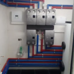 Instalacja grzewcza z rurami w kolorze czerwonym i niebieskim, podłączona do srebrnych modułów na ścianie w pomieszczeniu z czarną podłogą.