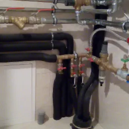 Złożony układ rur miedzianych i czarnych izolowanych, z licznymi zaworami i przyłączami, zamontowany przy ścianie w pomieszczeniu z kratką ściekową w podłodze.