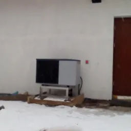 Zewnętrzna jednostka pompy ciepła typu powietrze-woda, zainstalowana przy białej ścianie budynku w zimowych warunkach, z widocznym opadem śniegu.