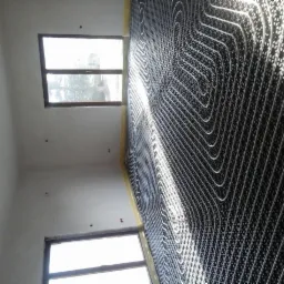 Białe rury ogrzewania podłogowego ułożone na czarnej macie izolacyjnej w nowo budowanym pomieszczeniu, widoczne okna i surowy stan ścian.