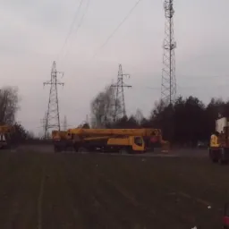 Żółte dźwigi i koparka na placu budowy, w tle słupy wysokiego napięcia i wieża telekomunikacyjna, pochmurne niebo. Sprzęt budowlany na trawiastym terenie.