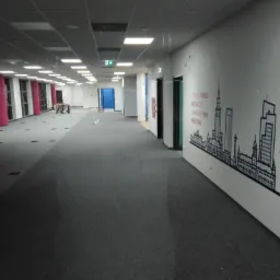 Długi korytarz biurowy z szarą wykładziną, białymi ścianami ozdobionymi grafiką przedstawiającą panoramę miasta i logo firmy, widoczne drzwi i okna, sufit podwieszany z oświetleniem.