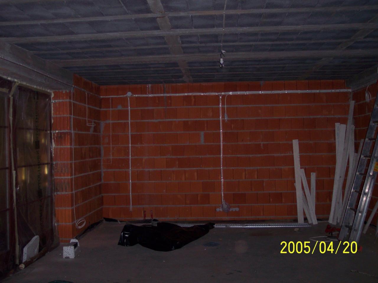 Surowy mur z przewodami elektrycznymi w peszlach, widoczne kable i puszki. Na podłodze pozostałości materiałów budowlanych. W tle drabina i elementy konstrukcyjne.