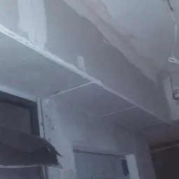 Karton-gips na suficie i ścianach w trakcie montażu. Widoczne łączenia płyt i przewody elektryczne. Ujęcie z dołu, widać fragment okna.