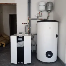 Biało-czarna jednostka sterująca z dwoma wyświetlaczami temperatury obok białego zbiornika akumulacyjnego z manometrem, szare rury instalacyjne z zaworami i dwa białe zbiorniki wyrównawcze...