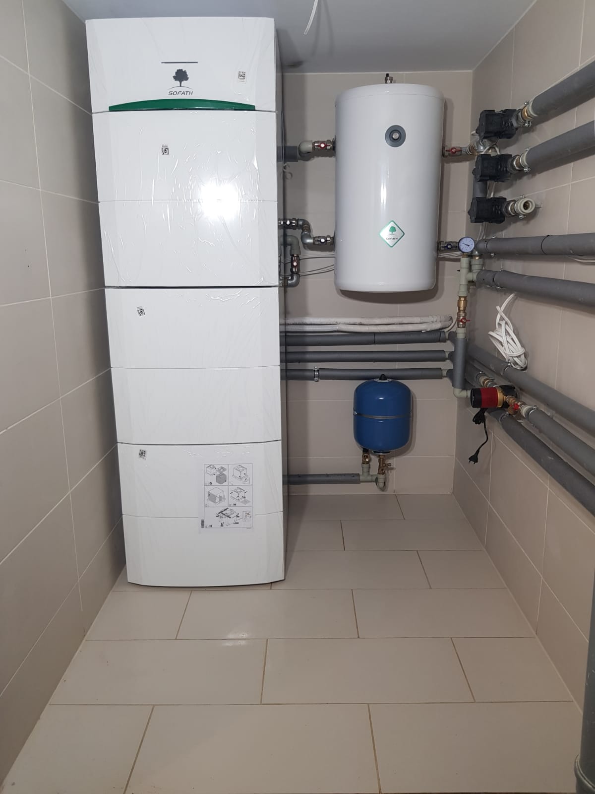 Nowa, jeszcze ofoliowana instalacja grzewcza Sofath, z białym zbiornikiem akumulacyjnym, niebieskim naczyniem przeponowym i szarymi rurami w pomieszczeniu wyłożonym jasnymi płytkami.