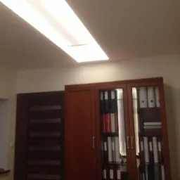 Wnętrze pokoju z wbudowaną szafą z przeszklonymi drzwiami, widoczna lampa sufitowa i drzwi z poziomymi przeszkleniami.