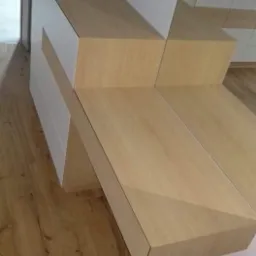 Boczny widok schod&oacute;w wewnętrznych z jasnego drewna, wbudowanych w szafę z białymi frontami szuflad, na drewnianej podłodze.