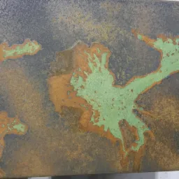 Zbliżenie na fragment blatu z nieregularnymi, zielonymi i rdzawymi plamami na ciemnym tle, przypominające mapę lub abstrakcyjny wzór.