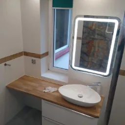 Nowoczesna łazienka po renowacji: białe płytki, drewniany blat z umywalką nablatową, podświetlane lustro, szare płytki podłogowe i okno z zieloną roletą.