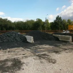 Góra ekogroszku na placu składowym, oddzielona betonowymi barierami. W tle drzewa i błękitne niebo z chmurami. Widok z poziomu ziemi.