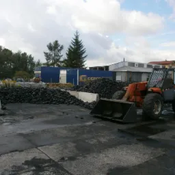 Pomarańczowa ładowarka teleskopowa przed stertą antracytu na placu składowym, w tle budynki i niebieski kontener z logo firmy CEFCO.