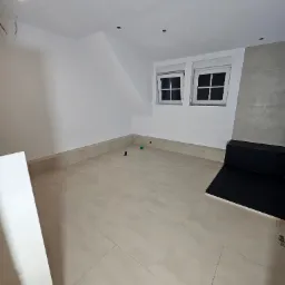 Jasne, świeżo ułożone płytki podłogowe w pokoju na poddaszu, z niską ścianką z płytek przy ścianie. Dwa okna dachowe w białych ramach, szara ściana z płytek.