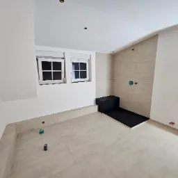 Jasna łazienka w trakcie wykańczania z beżowymi płytkami, czarnym brodzikiem i dwoma oknami. Widoczne niedokończone instalacje i punkty świetlne w suficie.