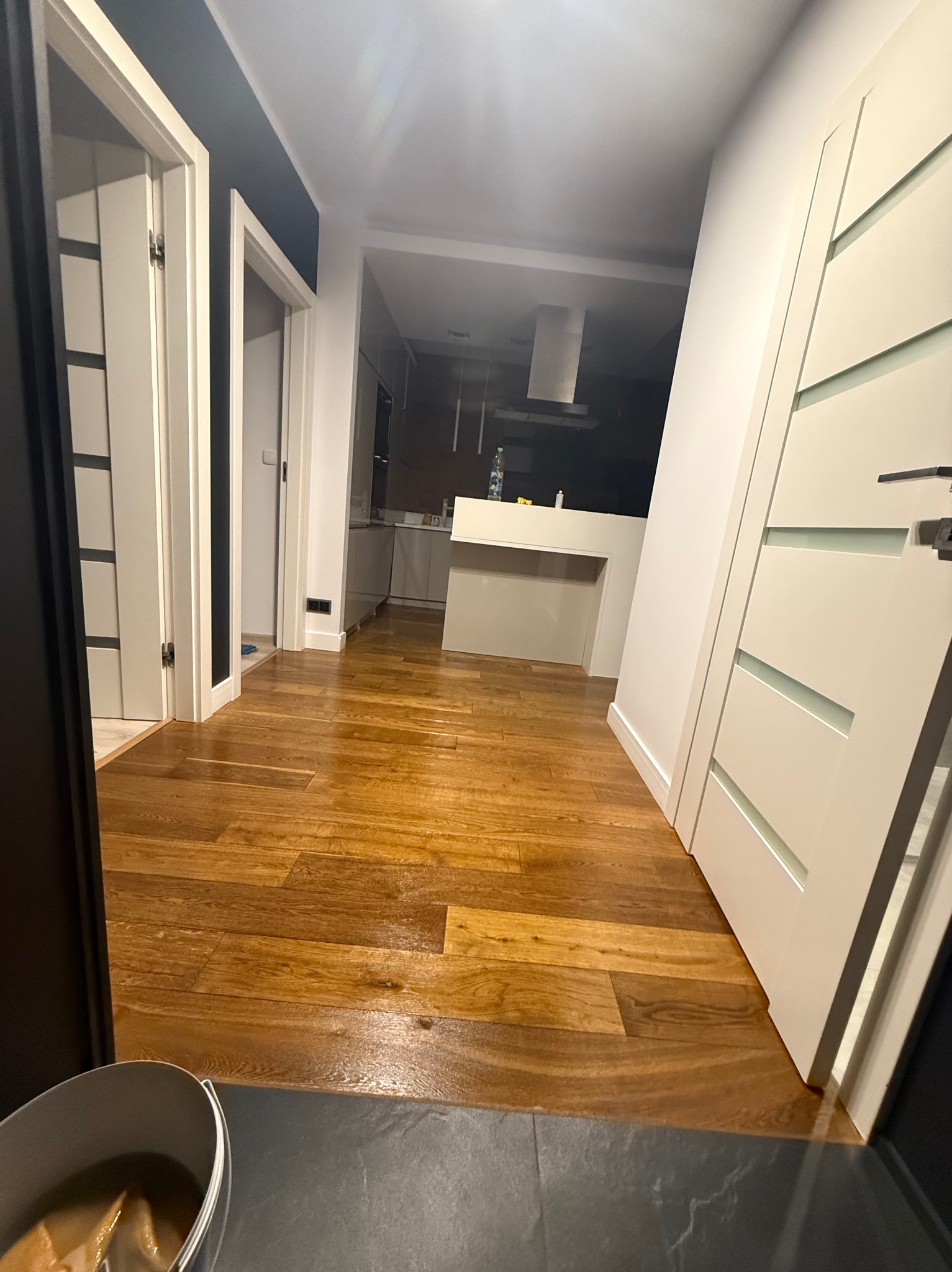 Błyszcząca, świeżo umyta podłoga parkietowa w korytarzu, z widokiem na kuchnię. Widoczne białe drzwi i wiadro z mopem w rogu kadru.