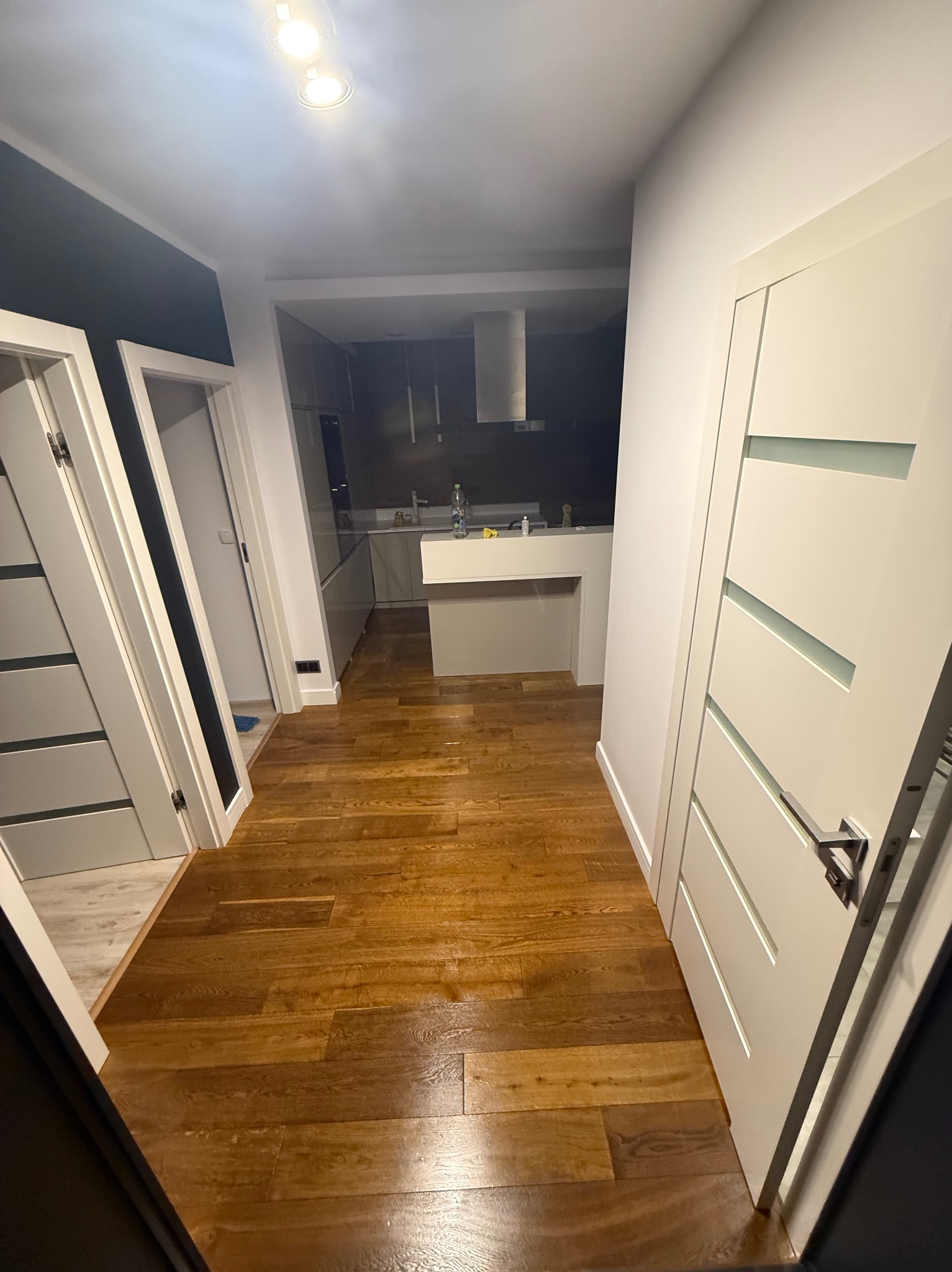 Ułożony parkiet w odcieniach brązu, widok z korytarza na kuchnię w Polance Wielkiej. Białe drzwi z przeszkleniami po bokach kadru.