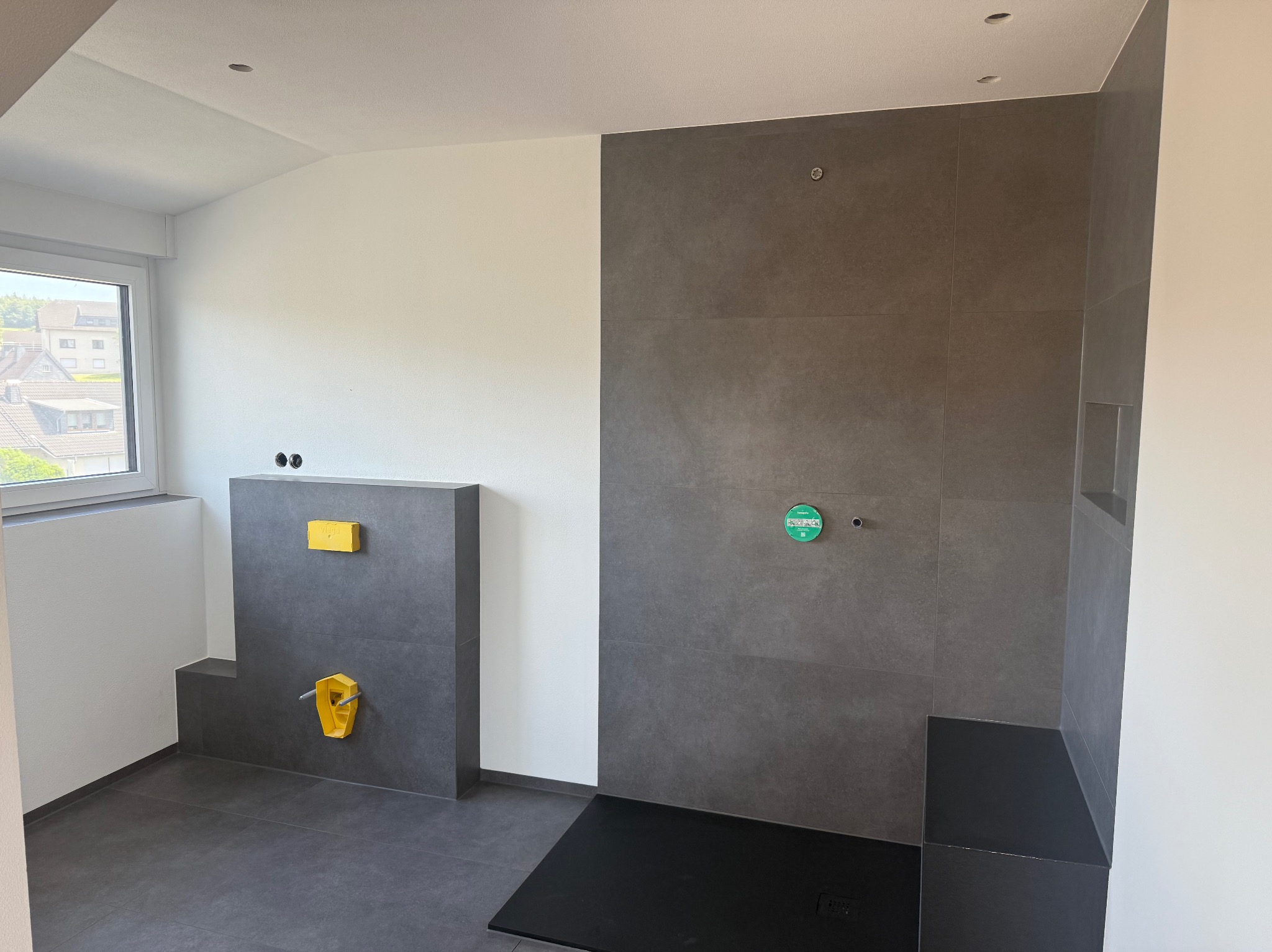 Szara, minimalistyczna łazienka w trakcie wykańczania, z widoczną zabudową WC i prysznicem typu walk-in z czarnym brodzikiem. Widoczne okno i surowe ściany.