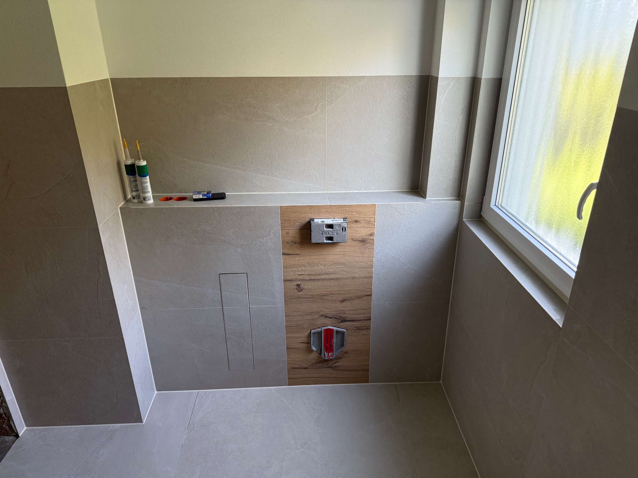 Wykończona łazienka z beżowymi płytkami, drewnopodobną wstawką z przyciskami spłuczki i oknem. Widoczne silikony i narzędzia glazurnicze.
