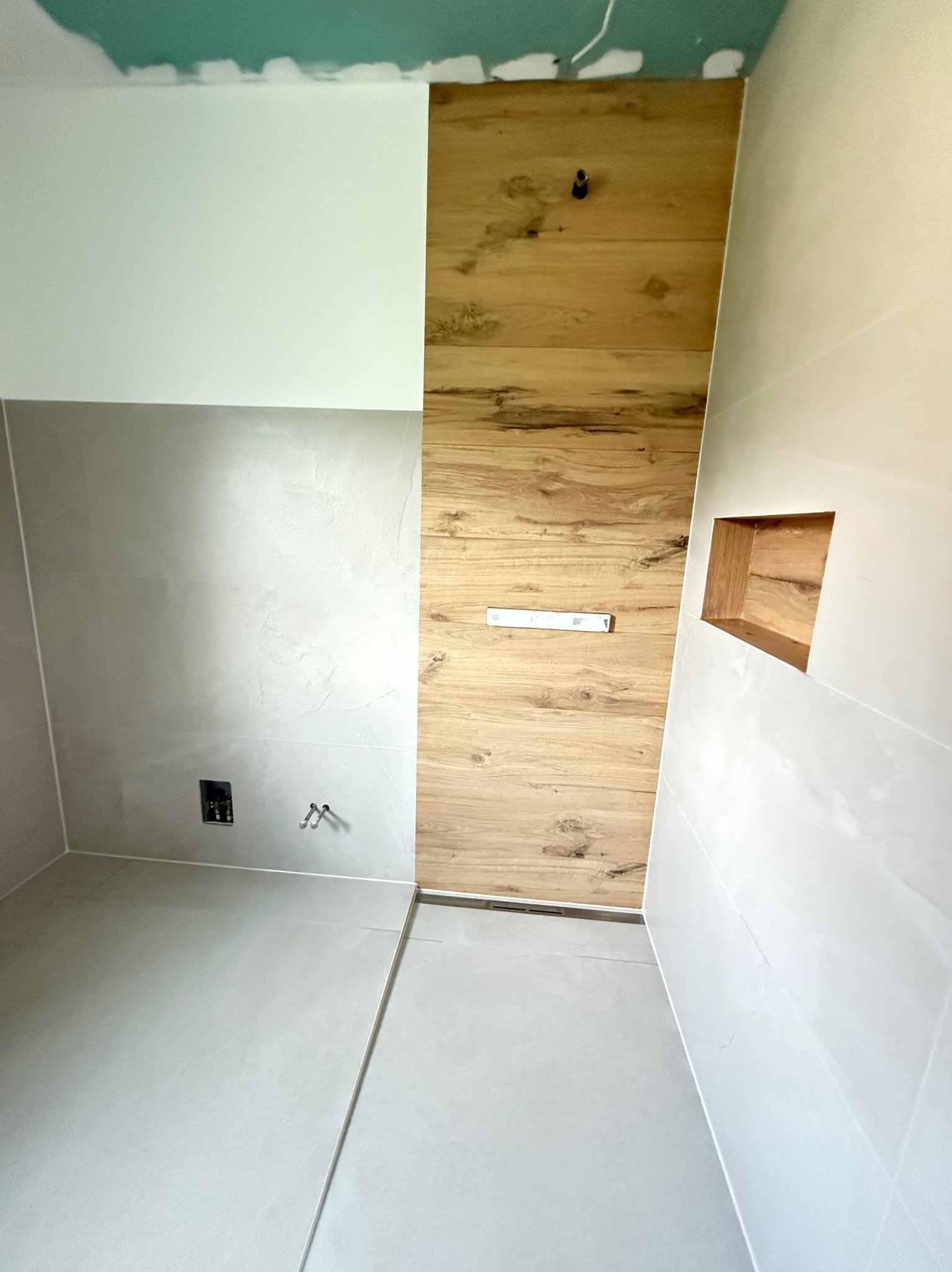 Wykończona łazienka z drewnopodobną ścianą, półką w ścianie, szarą podłogą i białymi płytkami. Widoczne przyłącza. Górna część ściany pomalowana na zielono.