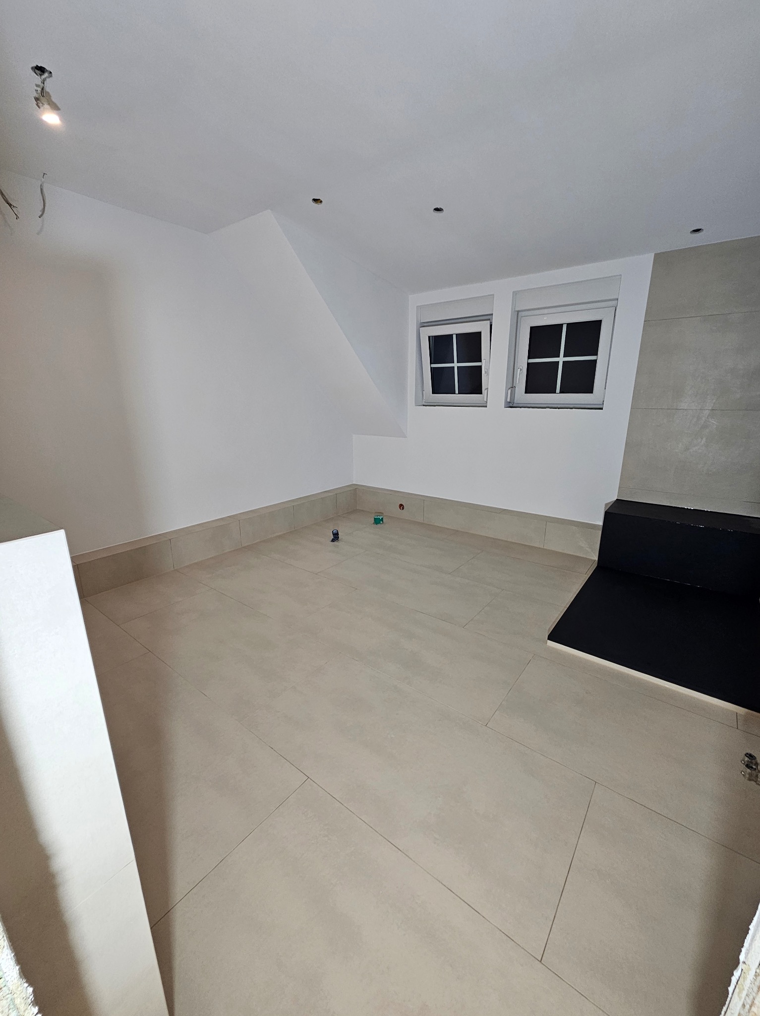 Jasne, świeżo ułożone płytki podłogowe w pokoju na poddaszu, z niską ścianką z płytek przy ścianie. Dwa okna dachowe w białych ramach, szara ściana z płytek.