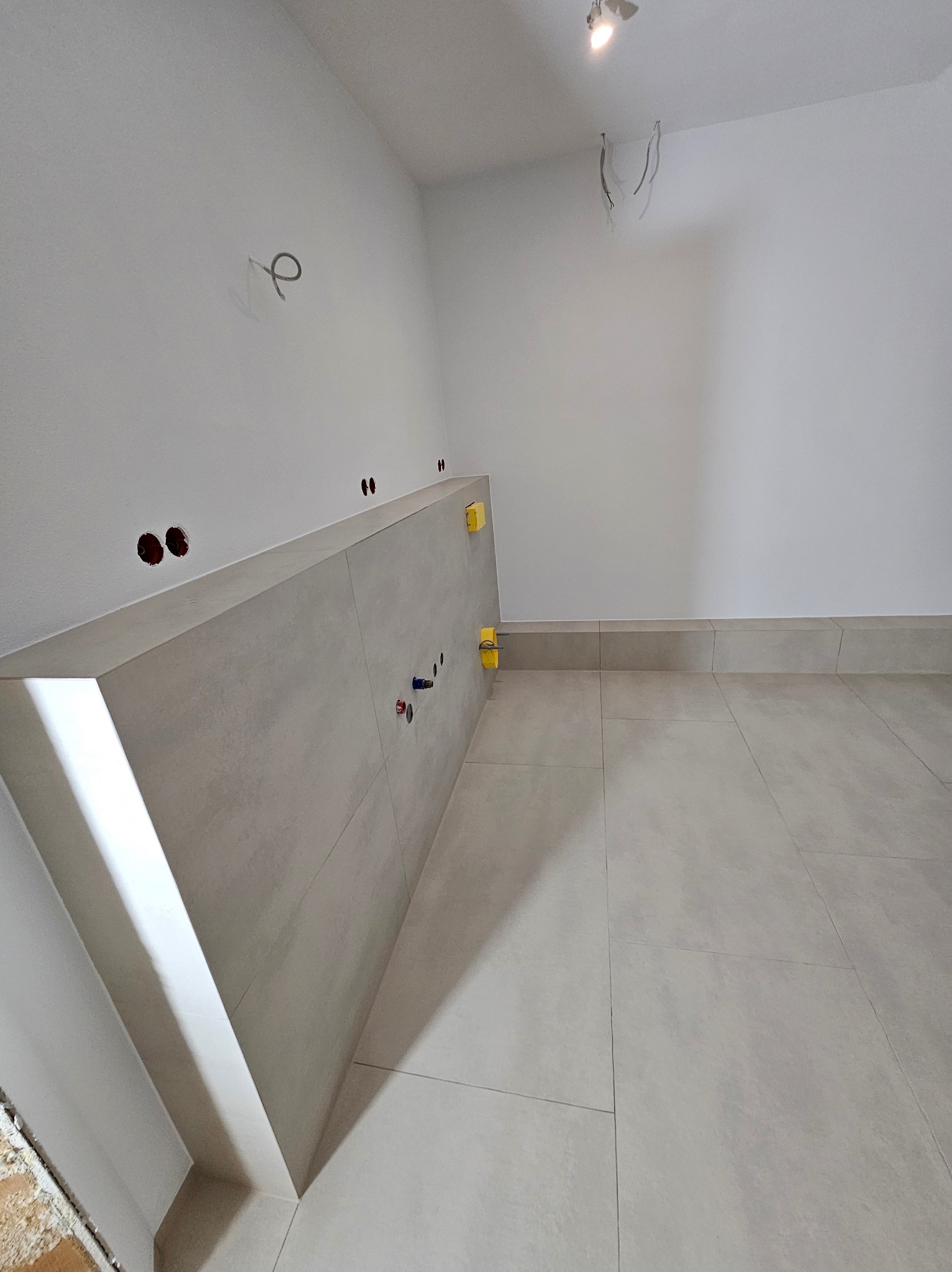 Jasne, matowe płytki w nowej łazience z częściowo wykończoną ścianką działową, widoczne otwory na instalacje i żółte kliny poziomujące płytki.