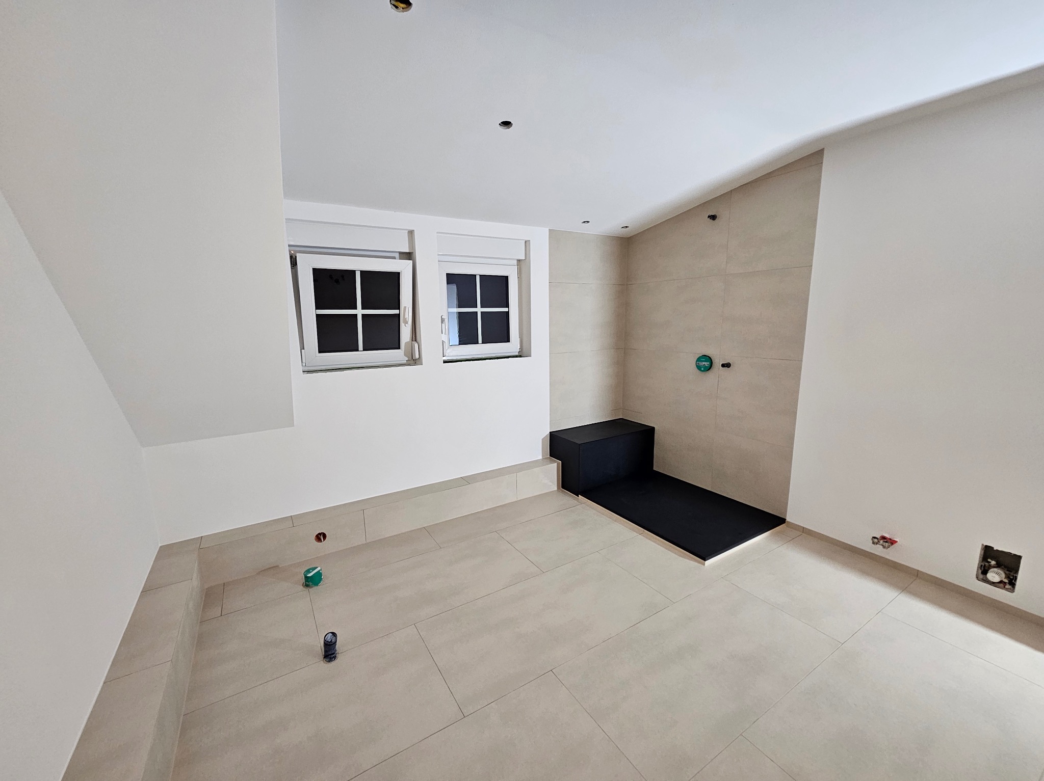 Jasna łazienka w trakcie wykańczania z beżowymi płytkami, czarnym brodzikiem i dwoma oknami. Widoczne niedokończone instalacje i punkty świetlne w suficie.